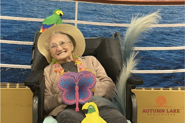 A smiling senior resident in a wheelchair wearing a sun hat and lei during a tropical-themed nursing home event.
