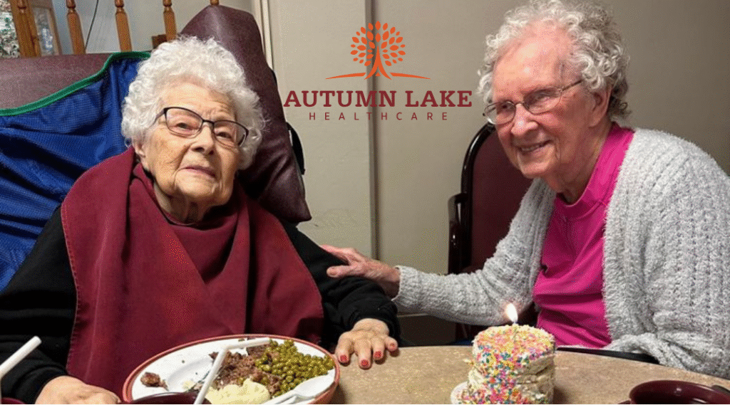Two elderly women smiling together at a nursing home table with a small birthday cake.